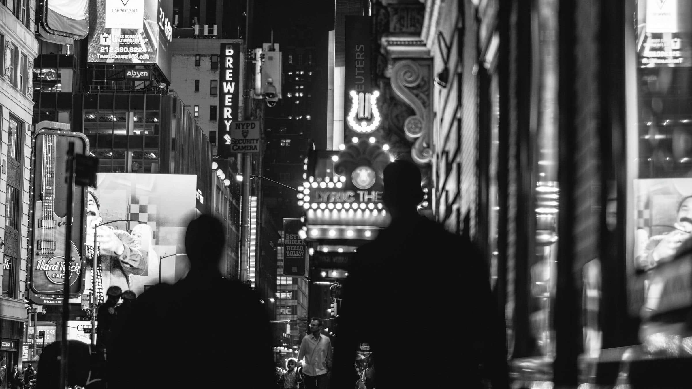Times Square at night in black and white, silhouettes walking beneath lit billboards